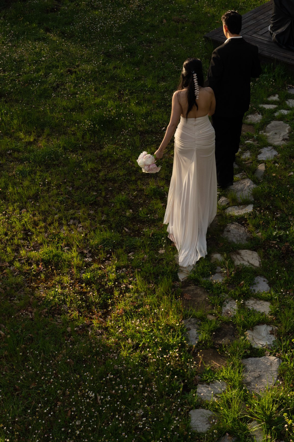Steph and Saw walking away along a stone path through spring grass at golden hour, her bouquet held at her side.