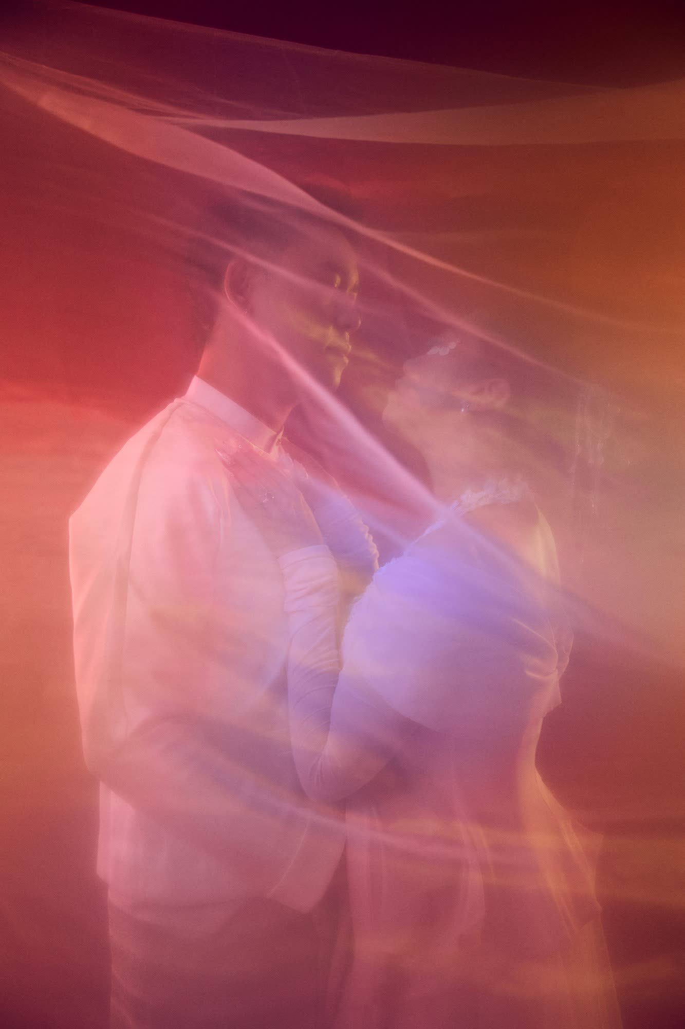 Steph and Saw holding each other beneath a long veil, lit in warm rose and amber.